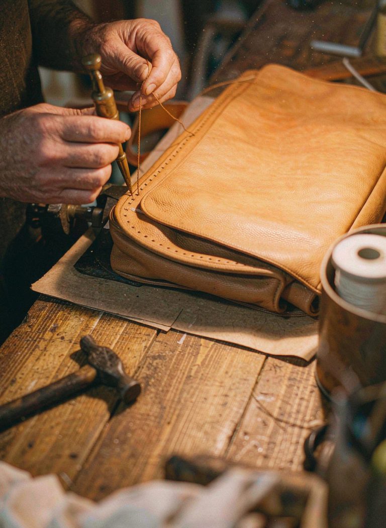 Craftsman hand-stitching the edge of a tan leather briefcase in a workshop
