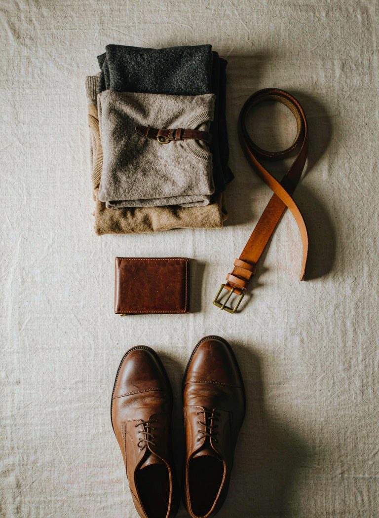 Flat lay of folded knitwear, a leather belt, card holder and derby shoes on linen